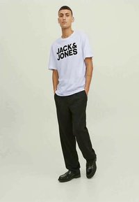 T-shirt de algodão branco com texto preto "JACK & JONES", combinada com calças pretas e sapatos de couro pretos. Design minimalista com corte descontraído.