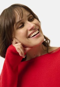 Maglione rosso lavorato a maglia con design off-the-shoulder, texture a coste e maniche lunghe. Il modello ha capelli castani di lunghezza spalla.