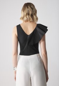 Svart bodysuit med djup V-rygg och enaxlad design med volangdetalj. Parat med högmidjade vita byxor.