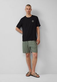 Zwarte katoenen t-shirt met een logo, lichtgroene shorts met een gestructureerde afwerking, en bruine instapsandalen met een decoratieve gesp.