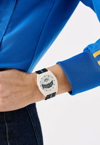 Montre ronde avec un cadran blanc et des graphismes bleus, bracelet noir avec des accents bleus, portée au poignet aux côtés d'une chemise bleue.