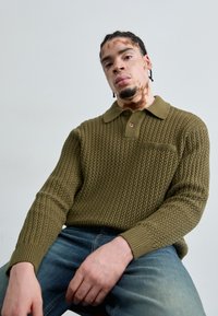 Khaki green knitted pullover with a collared neckline, textured pattern, and ribbed cuffs. Features a small chest pocket and button detail.