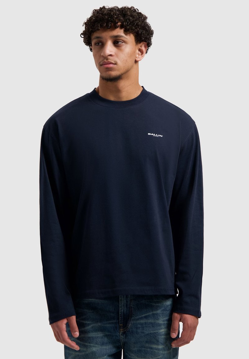 Langärmeliges Marineblaues T-Shirt aus weicher Baumwolle mit Rundhalsausschnitt und kleinem weißem Logo auf der linken Brust. Kombiniert mit hellblauen Jeans.