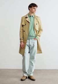 Jeune homme debout à l'intérieur portant un trench-coat beige, un polo vert clair, un jean bleu clair et des chaussures en daim marron clair.