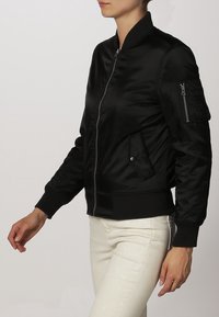 Veste bomber noire à finition brillante, avec une fermeture éclair devant, des poignets côtelés et une poche latérale. Fabriquée en tissu léger et lisse.