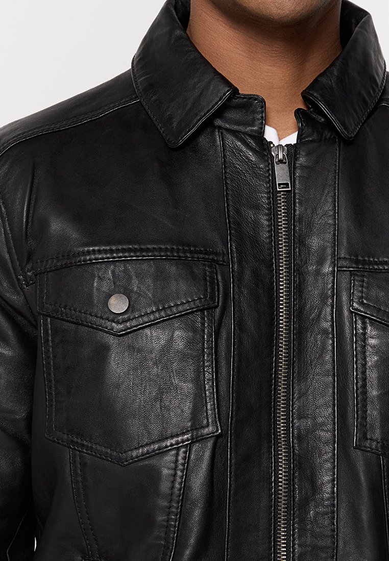Primo piano di una giacca di pelle nera con zip argentata, tasche petto a pattina con bottoni a scatto e colletto a punta su una persona che indossa una camicia bianca.