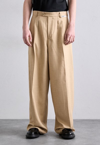 Beige wide-leg trousers with front pleats, made of a textured fabric. Two-button closure at the waist, paired with black shoes.