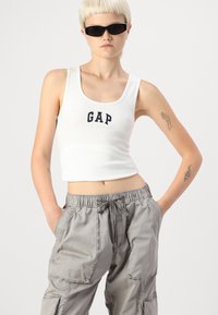 Un débardeur blanc côtelé avec le logo "GAP" en bleu marine, associé à un pantalon cargo léger gris clair avec poches et une taille élastique.