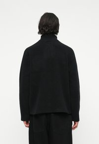 Jachetă neagră din fleece cu guler înalt și o suprafață texturată. Are umeri coboarăți și croială relaxată, combinată cu pantaloni negri.