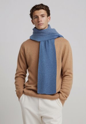 Sciarpa azzurro chiaro a coste realizzata in materiale morbido, drappeggiata intorno al collo. Indossata con un maglione color cammello e pantaloni color crema.