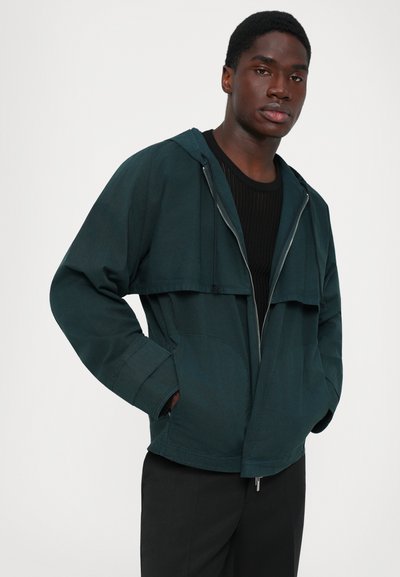 Νεαρός άνδρας φοράει μια σκούρα πράσινη ζακέτα με φερμουάρ και κουκούλα πάνω από ένα μαύρο πουκάμισο, με τα χέρια στις τσέπες, σε απλή ανοιχτόχρωμη φόντο.