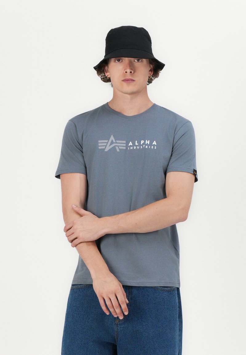 Graues kurzärmeliges T-Shirt mit einem Logo auf der Brust und einem runden Halsausschnitt, kombiniert mit blauen Jeans und einem schwarzen Bucket Hat.