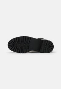 Semelle de botte noire avec des rainures profondes et larges, et des grips en forme de losange, présentée sur un fond blanc uni.