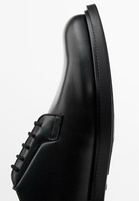 Black leather shoe with a smooth finish, rounded toe, and six lace-up eyelets. Distinct stitching along the side and a flat sole.