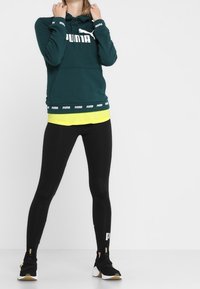 Tyrkysová mikina s kapucí a bílým logem, vrstvená přes žlutý top. Černé legíny a boty se zlatými detaily doplňují celý outfit.