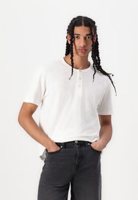 White short-sleeve henley shirt with three-button placket, textured fabric, paired with high-waisted black jeans.