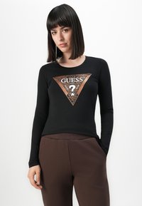 Donna con capelli lisci e scuri, che indossa una maglietta nera a maniche lunghe con un logo Guess color rame e pantaloni marroni, in piedi su uno sfondo bianco.