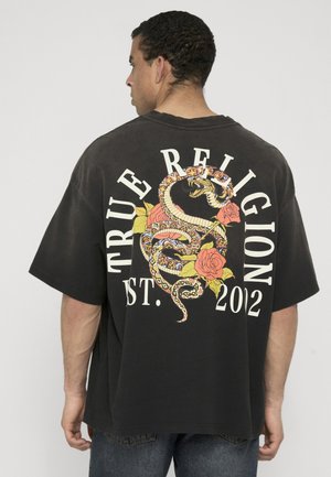 Man met zijn rug naar ons toe draagt een zwarte oversized blouse met een kleurrijk slang- en rozenontwerp en de tekst "TRUE RELIGION EST. 202" op de rug.
