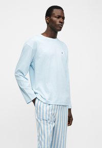 Άνδρας που φοράει ανοιχτό γαλάζιο πουκάμισο με μακριά μανίκια και λευκό παντελόνι με κάθετες ανοιχτό γαλάζιες ρίγες, στέκεται με το ένα χέρι στην τσέπη πάνω σε λευκό φόντο.