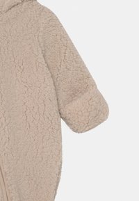 Veste en polar beige avec une surface douce et texturée, dotée d'une capuche et d'une fermeture éclair à l'avant. Design à manches courtes.
