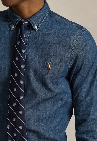 Uomo che indossa una camicia di jeans blu con bottoni con logo di giocatore di polo e una cravatta a righe blu navy con motivi di stemma.