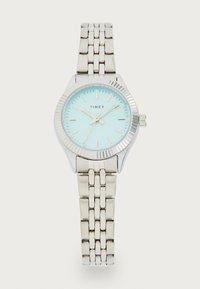 Montre Timex argentée avec un cadran bleu clair et un bracelet en argent. Elle présente une lunette texturée, des index horaires et trois aiguilles pour l'affichage de l'heure.