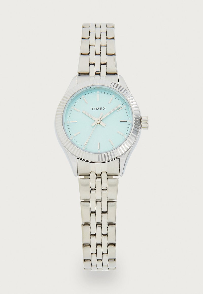 Montre Timex argentée avec un cadran bleu clair et un bracelet en argent. Elle présente une lunette texturée, des index horaires et trois aiguilles pour l'affichage de l'heure.