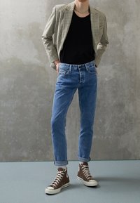 Kostkovaný blazer v béžové a zelené barvě přes černé tričko, doplněný o světle modré džíny a hnědé vysoké tenisky.