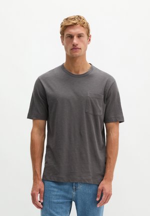 Uomo con capelli biondi corti che indossa una t-shirt grigio scuro a maniche corte con tasca e jeans blu, in piedi davanti a uno sfondo bianco semplice.