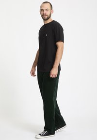 Zwarte katoenen T-shirt met korte mouwen en een klein logo op de borst, gecombineerd met donkergroene ribfluweel broek en zwarte sneakers.