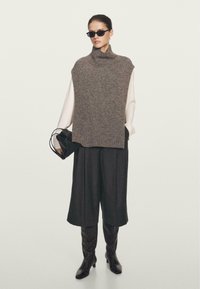 Grijze gebreide mouwloze coltrui, gecombineerd met een wit shirt met lange mouwen, donkere wijde broek en laarzen tot de knie in het zwart. Zwarte handtas, zonnebril.