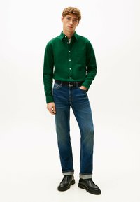 Groene button-up shirt met een borstzak, gecombineerd met blauwe spijkerbroek. Zwarte riem en enkellaarsjes. Effen achtergrond voor contrast.