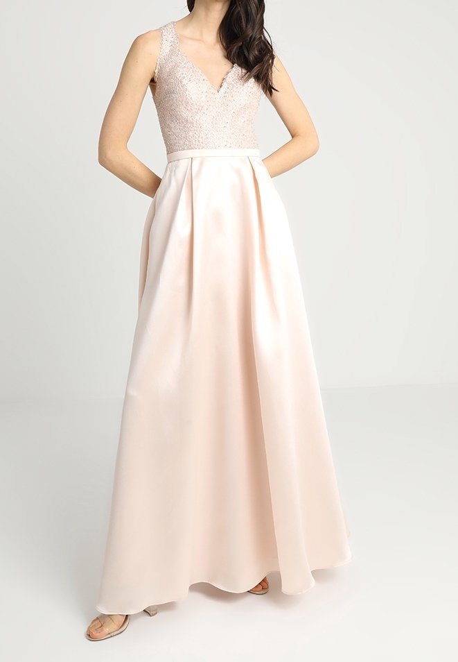 Femme portant une robe sans manches rose pâle avec un corsage en maille texturée en forme de V et une jupe lisse et fluide, debout, les mains dans le dos.