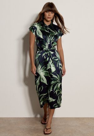 Donna con capelli castani lisci indossa un vestito blu navy con stampa di foglie verdi, maniche corte, vita annodata e sandali marroni, in piedi in casa.