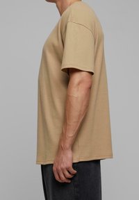 Vue de côté d'une personne portant une chemise beige à manches courtes texturée et un pantalon foncé sur un fond gris uni.