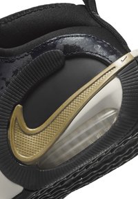 Sneaker nera con tomaia testurizzata, logo Nike dorato in evidenza e suola bianca con imbottitura e unità d'aria visibile. Suola in gomma con motivo antiscivolo.