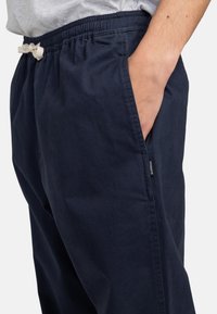 Pantalon en coton bleu marine avec une taille élastique, des poches latérales et une petite étiquette de marque sur le côté gauche. Coupe douce et décontractée.