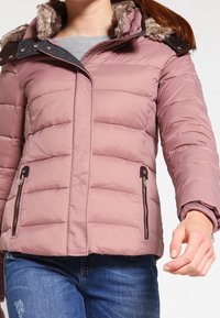 Puszysta, mauve kurtka z kapturem wyłożonym sztucznym futrem, z zamkniętymi kieszeniami po bokach i pikowanym wzorem. Posiada ciemne akcenty przy kołnierzu i zamkach.