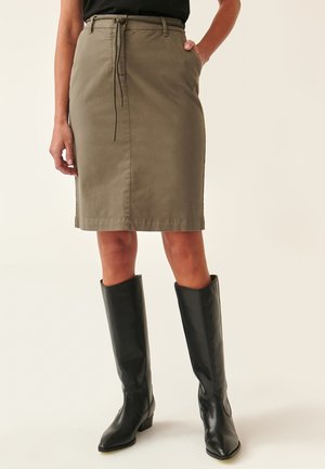 Jupe longueur genou vert olive avec taille à cordon, poches latérales et texture lisse, associée à des bottes en cuir noir hautes.