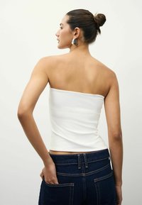 Biała, bezramiączkowa bluzka crop top wykonana z gładkiego materiału, zestawiona z ciemnoniebieskimi dżinsami z widocznymi szwami i tylnymi kieszeniami.