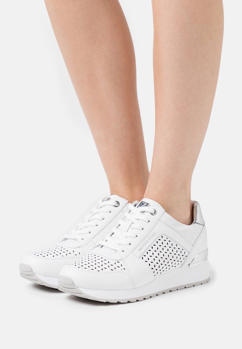 Paire de baskets blanches en cuir perforé avec lacets portées pieds nus sur un fond blanc uni.
