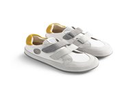 Sneakers bianchi con una punta in suede grigia, cinturini in Velcro grigi e un accento giallo sole sul tallone. Presentano un logo circolare sul lato.