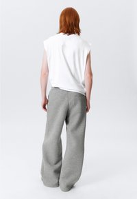Person med rødt hår står med ryggen til iført en ærmeløs hvid top og løse grå sweatpants foran en lys ensfarvet baggrund.