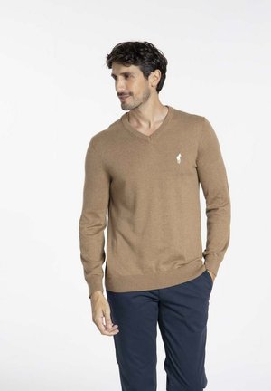 Pull en V marron avec une texture douce, arborant un petit logo blanc sur la poitrine gauche, associé à un pantalon bleu marine.