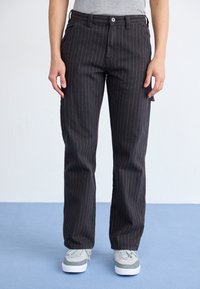 Svarta randiga byxor i slitstarkt tyg med rak bendesign och framfickor, tillsammans med grå sneakers.