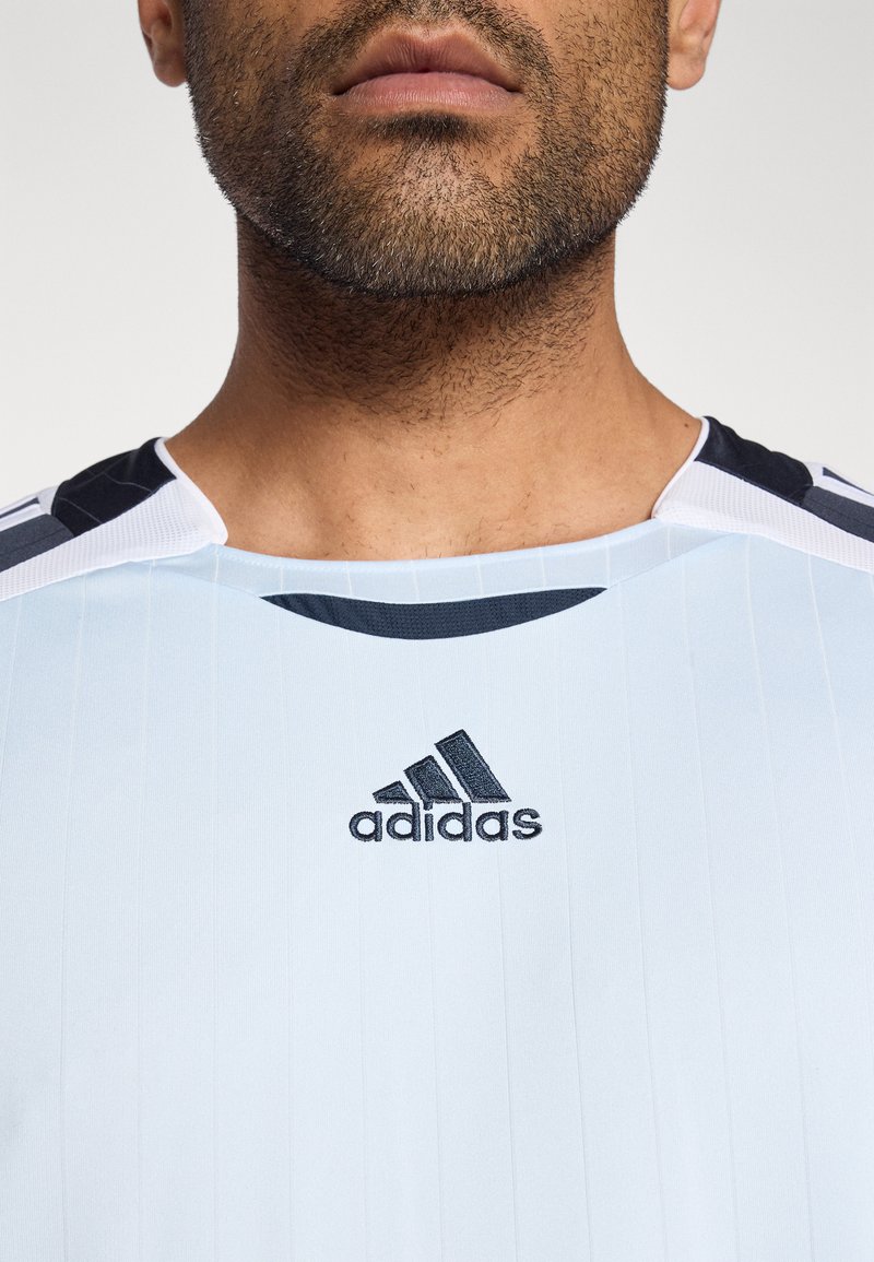 Maillot de sport bleu clair avec fines rayures horizontales, accents noirs sur les épaules et logo Adidas noir sur la poitrine. Texture lisse.