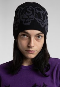 Beanie en tricot noir avec un design graphique gris foncé comportant des motifs de crânes. Le tissu semble lisse et ajusté, adapté aux temps frais.