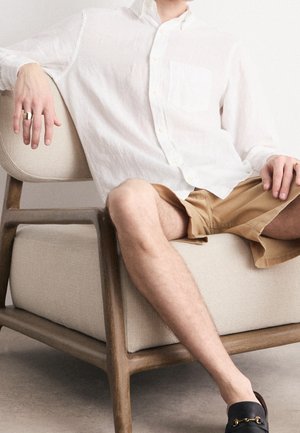 Personne portant une chemise blanche à boutons, un short beige et des chaussures noires sans lacets, assise sur une chaise en bois avec coussin beige.