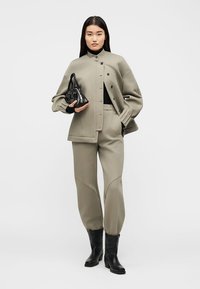 Ženska, oblečena v taupe oversized jakno in ujemajoče se hlače jogger, črni rolkasti pulover, črni gležnjarji, drži majhno črno torbico.