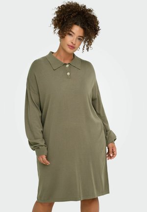 Femme aux cheveux bouclés portant une robe longue ample vert olive à manches longues avec un col et deux boutons, debout devant un fond clair uni.